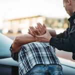 Police officer arresting person against car.