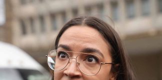 Person with glasses speaking outside, looking serious.