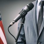 Man in suit speaking into microphone next to an American flag.