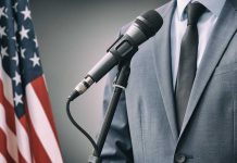 Man in suit speaking into microphone next to an American flag.