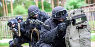 SWAT team members aiming rifles behind a protective shield.