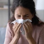 Hospital Chiefs Brace — 2025 Flu Outlook Darkens Woman sneezing into a tissue indoors.