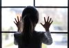 A child with long hair presses their hands against a window, looking outside