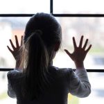 Father Forced Child Into Horrific Murder Cover-Up A child with long hair presses their hands against a window, looking outside