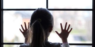 A child with long hair presses their hands against a window, looking outside