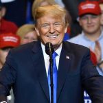 3rd Term Plot Thickens Man in suit speaking at rally with red hats.