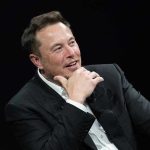 Musk Exposes Judge Bias Bomb Man in suit smiling, resting chin on hand.
