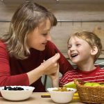 A mother playfully feeding her son in a kitchen filled with fruits and snacks