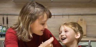 Junk Food REWIRES Kids’ Brains – Shocking Details! A mother playfully feeding her son in a kitchen filled with fruits and snacks