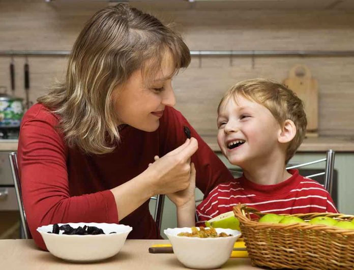 A mother playfully feeding her son in a kitchen filled with fruits and snacks