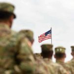 Soldiers in uniform standing, American flag in background.