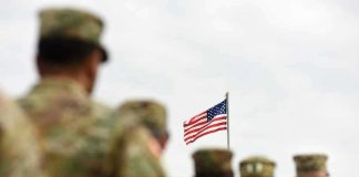 Soldiers in uniform standing, American flag in background.