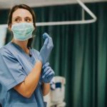 Nurse wearing scrubs and mask putting on gloves.