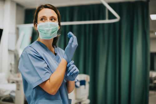 shutterstock1229815867-1jpg Nurse wearing scrubs and mask putting on gloves.
