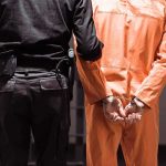 U.S. Marine Hospitalized — Russia Blocks Access Prisoner in orange jumpsuit handcuffed with guard standing beside