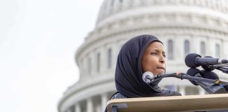 Woman in hijab speaking at podium with microphones outside a domed building