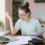 Young woman with glasses frustrated at laptop while holding bills