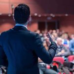 Viral Clip Resurfaces — Stakes Now Bigger Speaker addressing an audience in a conference hall