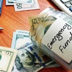 One Decision In 2008 Reshaped Global Finance Glass jar labeled emergency fund with US dollar bills on a wooden desk