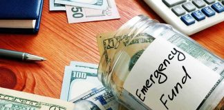 Glass jar labeled emergency fund with US dollar bills on a wooden desk