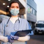 Healthcare worker in scrubs wearing mask and gloves holding a clipboard