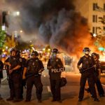Christian Jones and Teddy Williams among police line at night protest with fire and smoke