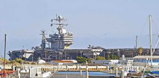 A U.S. Carrier Faces A 16 Month Repair Window A large naval aircraft carrier docked in a harbor with smaller boats in the foreground