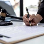 Hand signing legal document beside gavel and laptop
