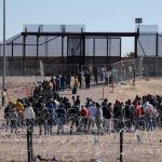 Crowd of people lining up near a border fence with barbed wire