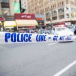 Police tape cordons off a city street with blurred buildings and patrol car