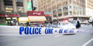 Police tape cordons off a city street with blurred buildings and patrol car