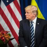 man in dark suit seated before US and Ukrainian flags