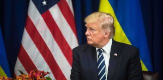 man in dark suit seated before US and Ukrainian flags