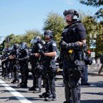 Bear Pascoe, Brian Folkerts, and Eugene Sims among riot police standing in a line on a city street