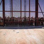 Crowd stands behind tall metal fence at border crossing